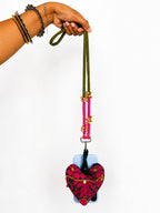 A hand wearing gold and silver bracelets holds a phone case with a long olive green strap, bright pink hanging cords, and a red and black patterned heart-shaped pouch with gold trim, against a plain white background.