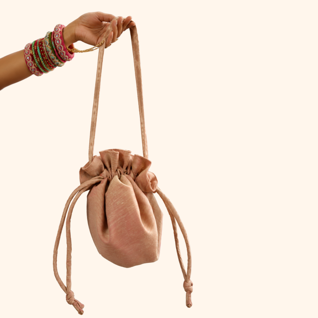 A blush pink velvet drawstring pouch bag held by a hand adorned with colorful bangles, set against a solid cream background.
