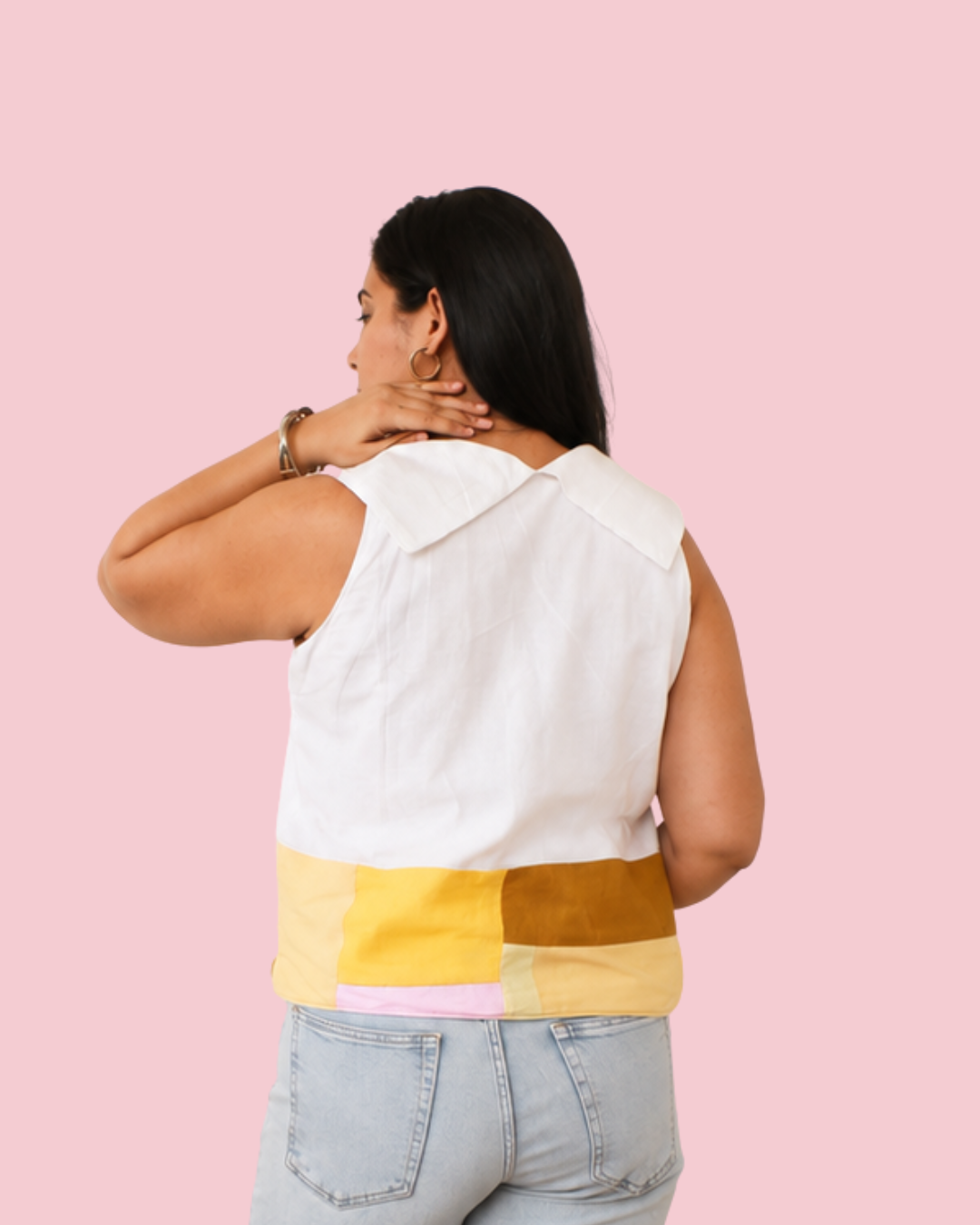 A woman shown from the back wearing a sleeveless white top with a wide collar and a color-blocked hem in yellow, mustard, and light pink, paired with light-wash jeans against a solid pink background.