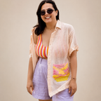 A woman modeling a light peach short-sleeve button-up shirt with a colorful abstract print on the front pocket, paired with lavender shorts and a striped tank top, against a solid beige background.