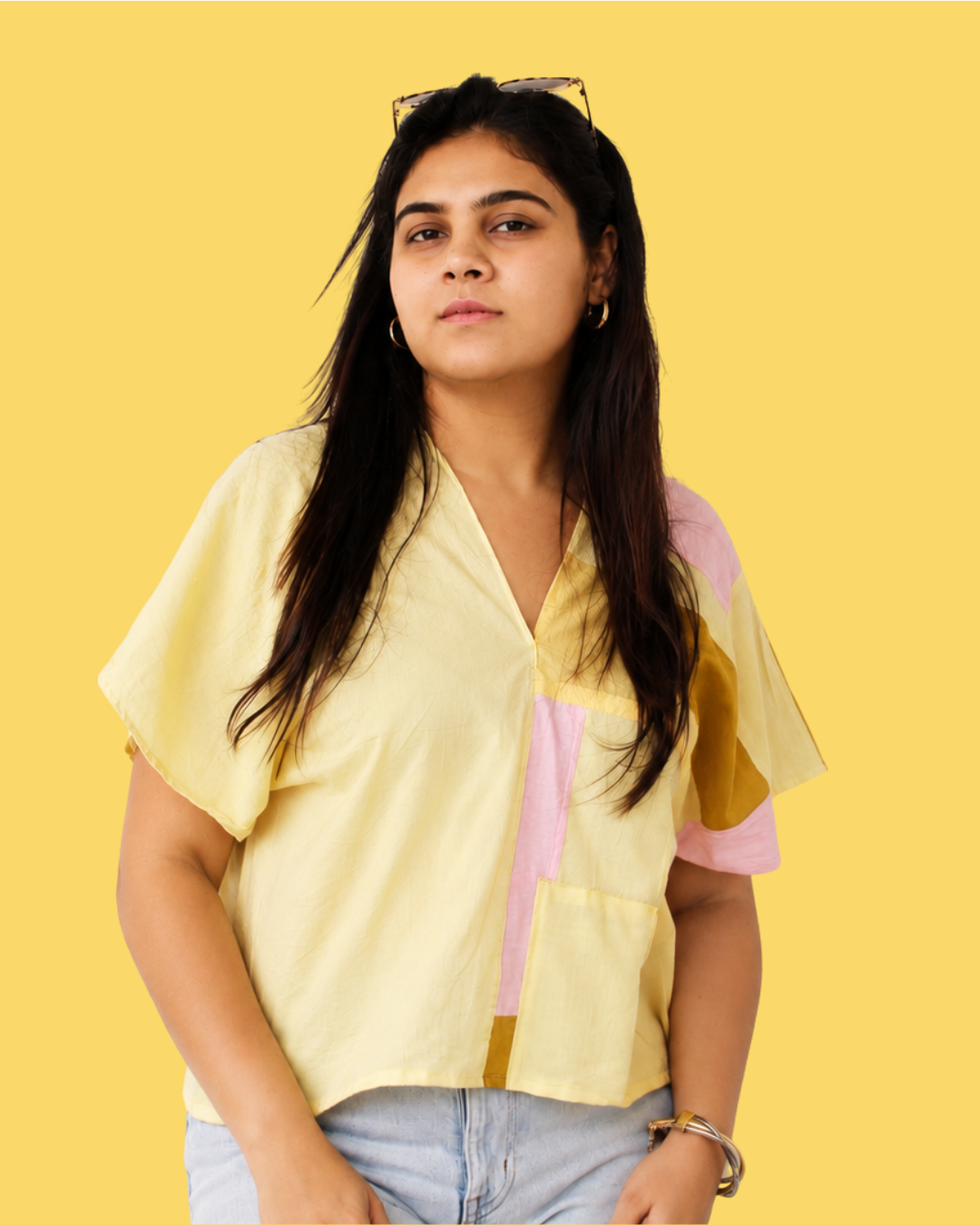 A woman modeling a short-sleeve, V-neck color-blocked blouse in pastel yellow, pink, and mustard yellow, paired with light blue jeans against a solid yellow background.