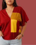 A woman wearing a loose-fitting short-sleeve top with a deep V-neck and large color-block panels in red and mustard yellow, paired with light blue jeans against a solid muted red background.