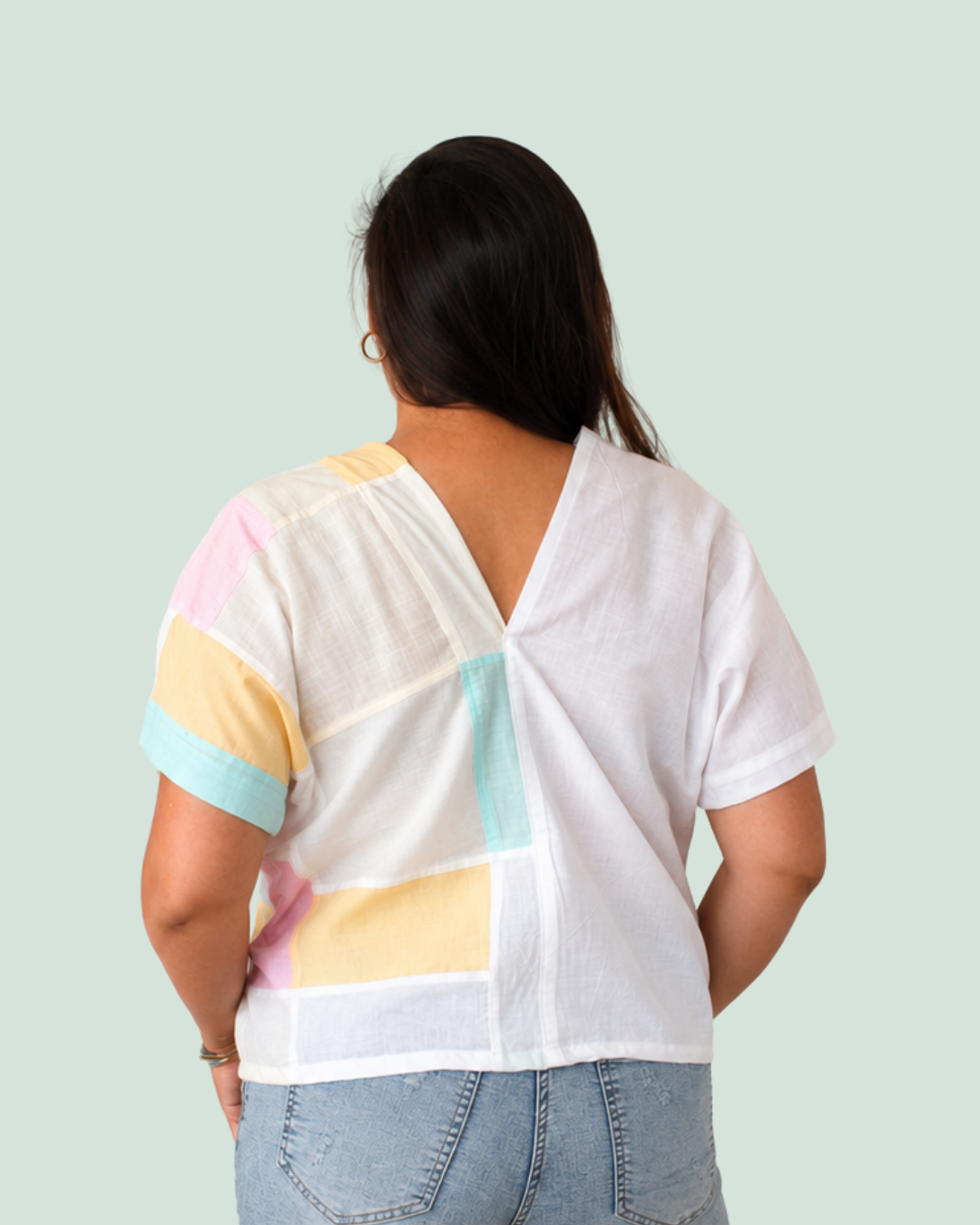 A woman viewed from behind wearing a white short-sleeve top with pastel geometric color-block panels in yellow, pink, and light blue, paired with light-wash jeans against a pale mint background.