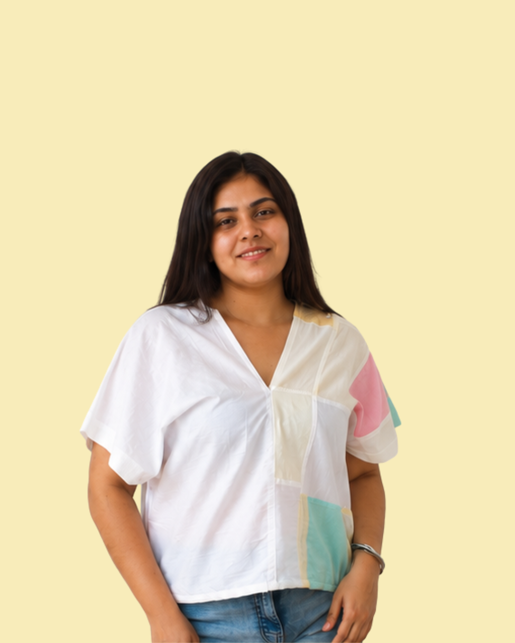 A woman wearing a white short-sleeve blouse with pastel color-block panels in pink, mint green, and cream, paired with blue jeans, set against a solid pale yellow background.