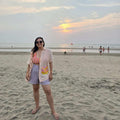 Woman standing on a beach at sunset with a colorful sky wearing a beach hand painted shirt