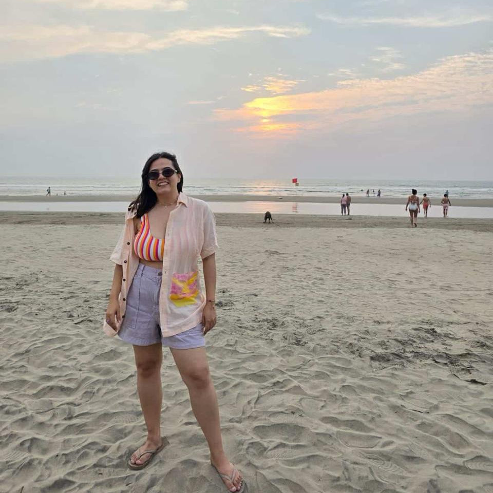Woman standing on a beach at sunset with a colorful sky wearing a beach hand painted shirt