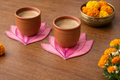 Two terracotta cups with pink lotus coasters on a wooden surface, surrounded by marigold flowers.