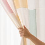 A hand holding back a sheer curtain panel with a patchwork design of pastel colors including cream, yellow, pink, mint green, and purple, set against a bright white window light.