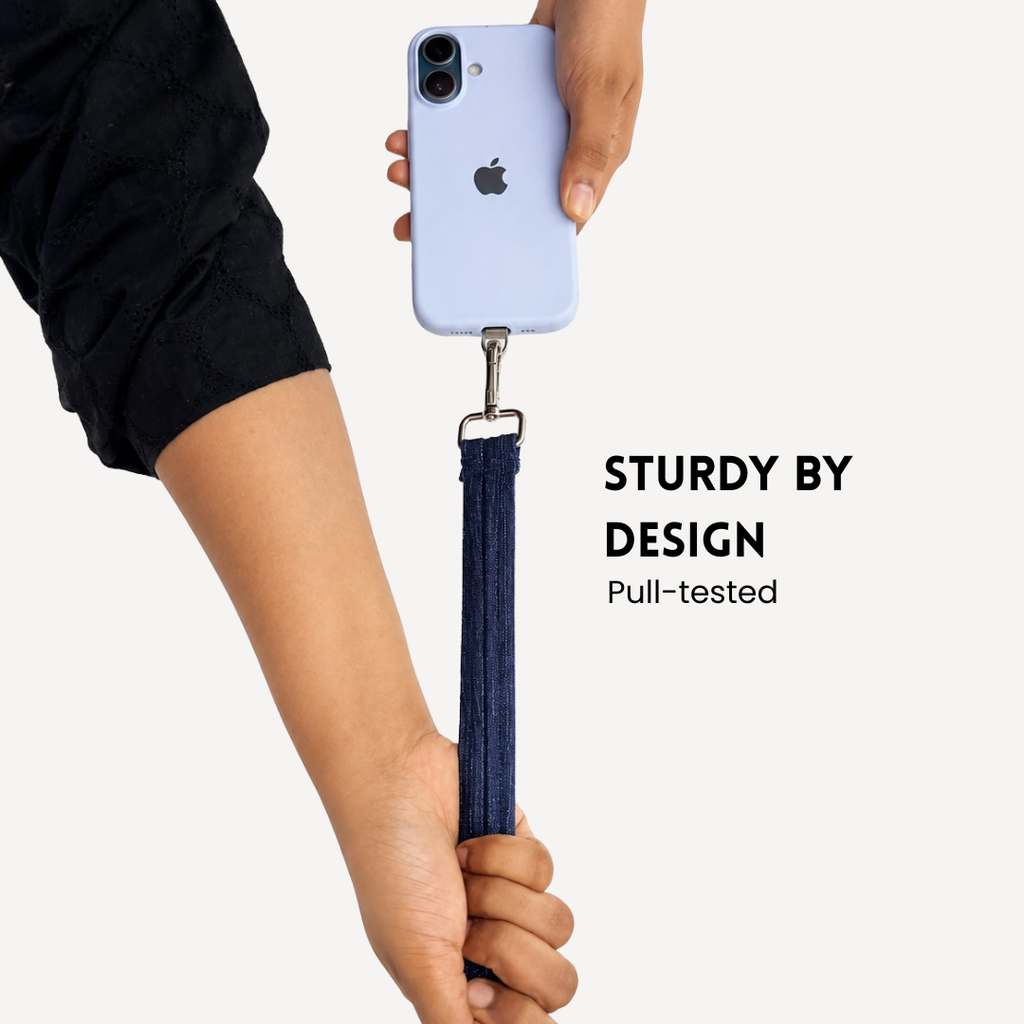 A person holds a light blue smartphone with a dark blue denim-style wrist strap attached by a metal clip. The Apple logo is visible on the phone, and the text reads 'STURDY BY DESIGN Pull-tested' on a light gray background.