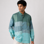 A man wearing a long-sleeve button-up shirt with a horizontal color-blocked pattern in shades of teal, green, and light blue, paired with off-white trousers, against a plain light gray background.