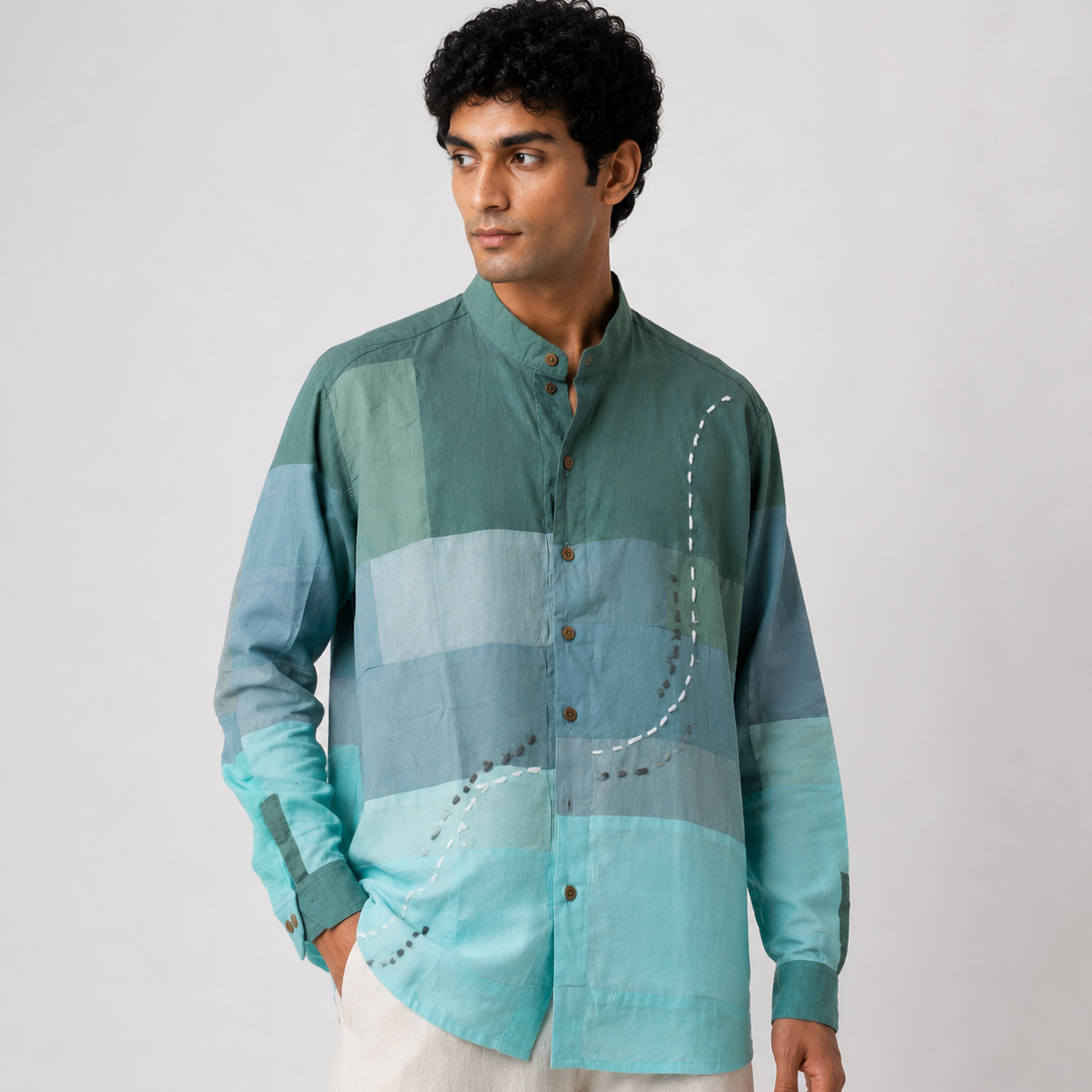 A man wearing a long-sleeve button-up shirt with a horizontal color-blocked pattern in shades of teal, green, and light blue, paired with off-white trousers, against a plain light gray background.