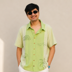 A man wearing a light green short-sleeve button-up shirt with embroidered tropical motifs, styled with white pants and dark sunglasses, standing against a plain beige wall.