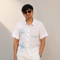 A man wearing a white short-sleeve button-up shirt with blue tropical embroidery, styled with white pants and dark sunglasses, standing against a plain beige wall.