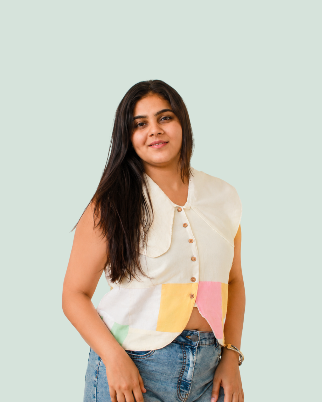 A woman wearing a sleeveless, asymmetrical button-front Vest top with pastel color-block panels in yellow, pink, and green on an off-white base, paired with blue jeans, set against a solid pale mint green background.