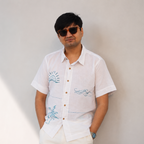 A man wearing a white short-sleeve button-up shirt with blue beach details on the chest, paired with white pants and dark sunglasses, standing against a plain light gray wall.