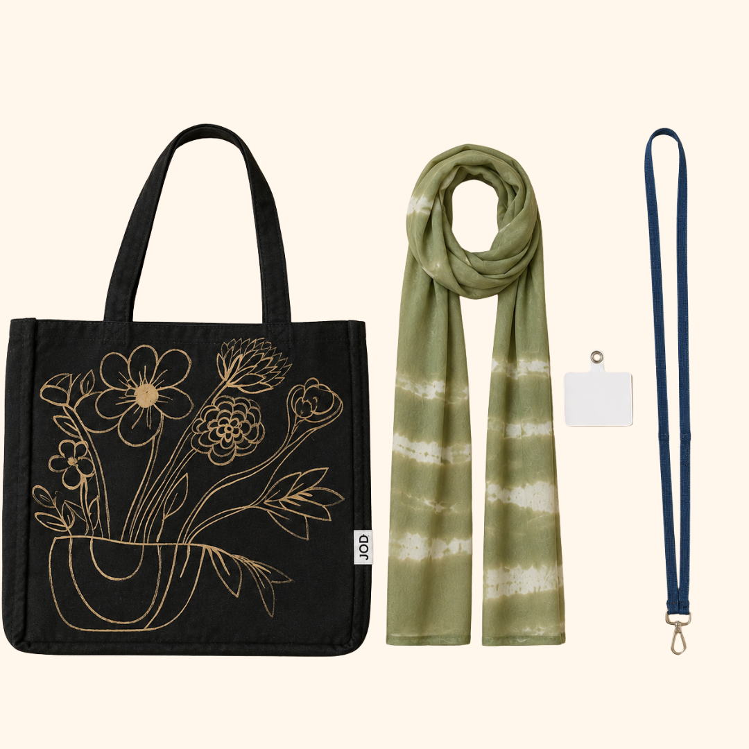 A flat-lay product set on a light cream background featuring a black canvas tote bag with a gold floral line art design, an olive green and white tie-dye scarf, a navy blue lanyard, and a small white card.