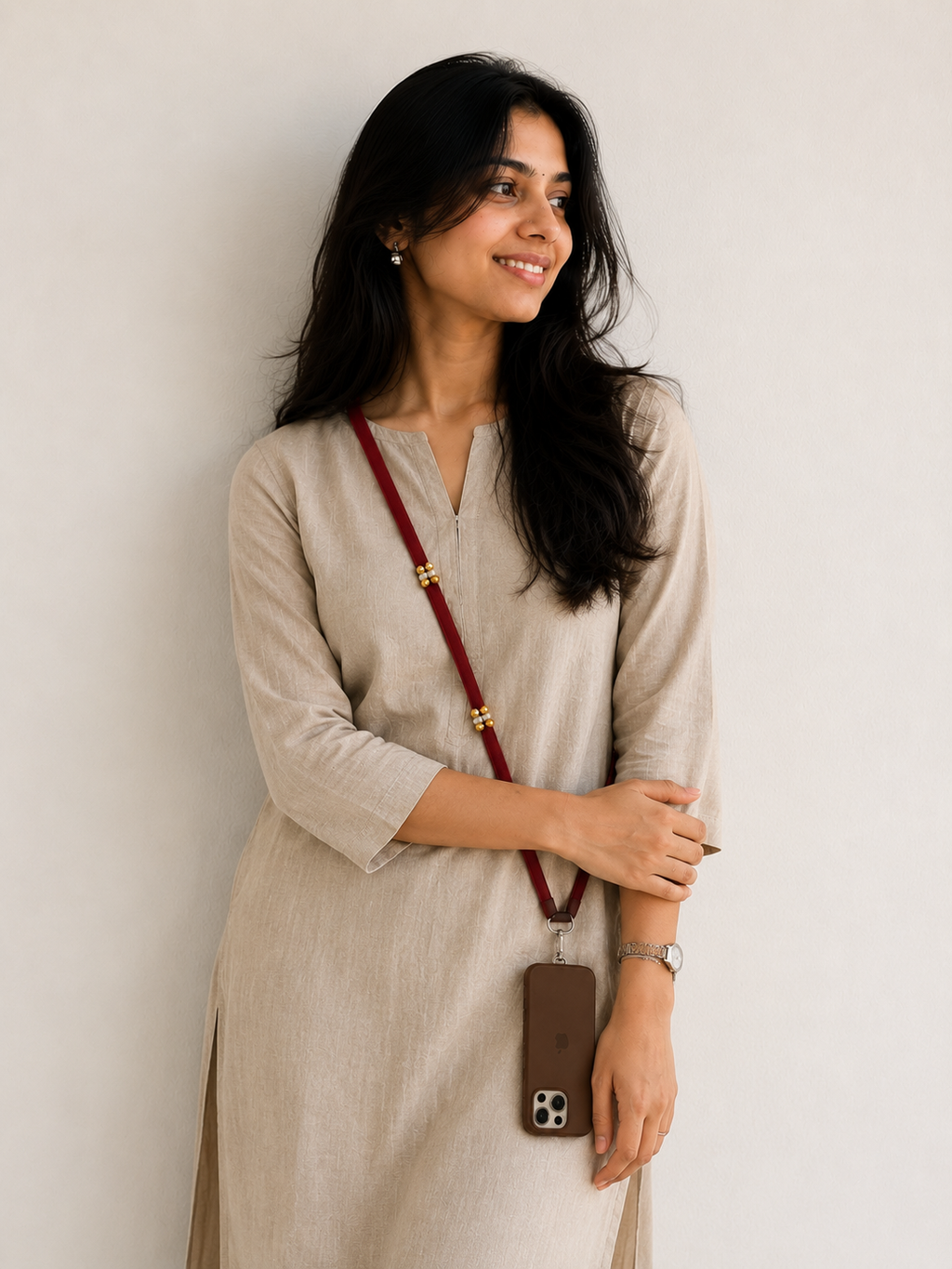 A woman wearing a light beige linen kurta with a split neckline and three-quarter sleeves, holding a brown leather phone case on a dark red crossbody strap with gold-toned decorative links.